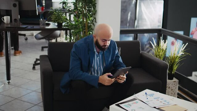 Employee comparing data on tablet while working in open space office. Unicorn business worker reviewing documents and annual statistics on device, trying to solve executive tasks.