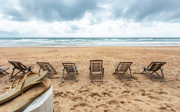 Espreguiçadeiras de madeira vazias na areia da praia deserta de frente para o mar em dia nublado. Chinelo desfocado em primeiro plano.