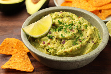 Bowl of delicious guacamole, lime and nachos chips on wooden table, closeup