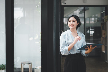 Happy young Asian saleswoman looking out the window welcoming client. Smiling woman executive manager, secretary offering professional business services holding digital tablet standing in office.