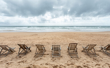 Fototapeta na wymiar Espreguiçadeiras de madeira vazias na areia da praia deserta de frente para o mar em dia nublado.