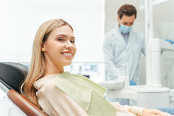 Young beautiful smiling woman, patient sitting in modern dental clinic with dentist on background. Dental treatment, health care concept