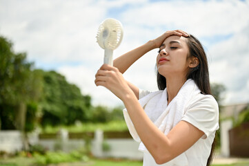 A sweaty Asian woman using a portable handy fan, feeling hot and tired after a long run