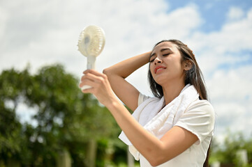 A sweaty Asian woman using a portable handy fan, feeling hot and tired, rests after a long run