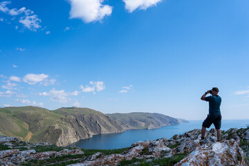 Fototapeta na wymiar person on the top of mountain. A young man is standing on a rock and looking at the sea through binoculars. Lake in summer. A tourist is resting on the shore and looking through binoculars. 