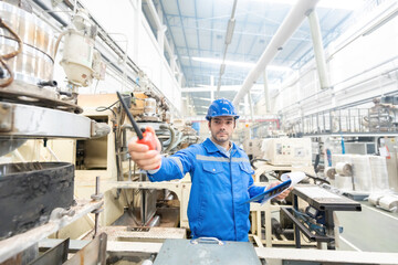 American male engineer, mechanic supervisor Put your hand in front of you holding a radio and a listnote. There is a working machine Wear a helmet and uniform. In the plastic and steel industry