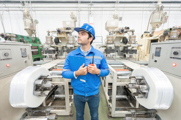 American male engineer, mechanic supervisor standing between machines look at the destination Holding a walkie-talkie, a lisnote, wearing a helmet and uniform. In the plastic and steel industry