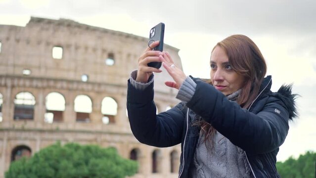 Happy young Latin woman tourist with luggage and warm clothes using smartphone in front of historic building Colosseum in Rome, Italy against cloudy sky