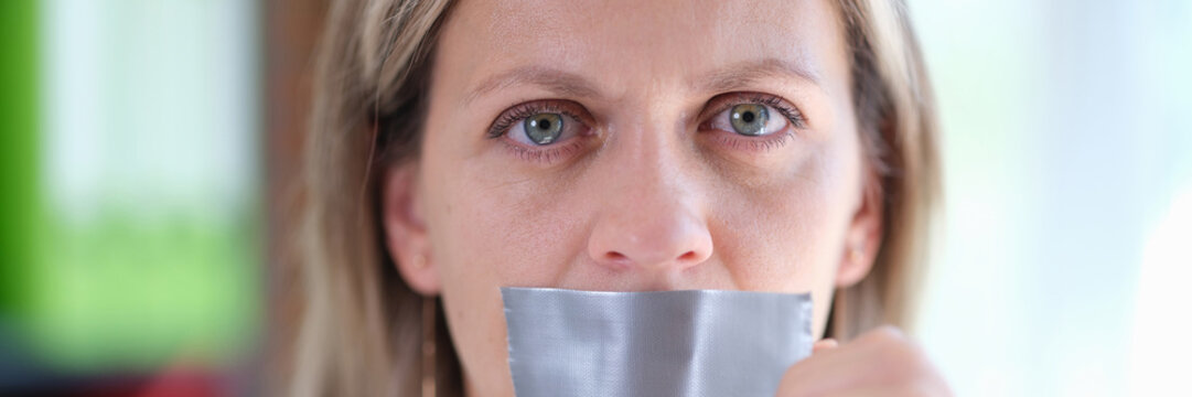 Female with serious face removes adhesive tape from her mouth close up.