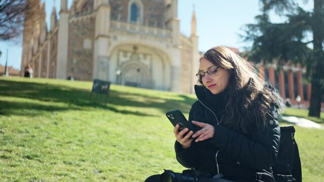 Young Latin woman tourist in warm clothes sitting on stone and using smartphone and professional photo camera while resting near church Saint Jerome the Royal next to Prado Museum in Madrid, Spain