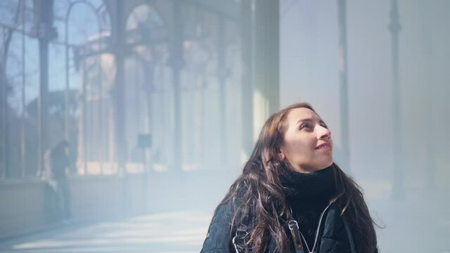 Happy young Latin woman tourist in warm clothes with professional photo camera walking through the mist and the rays of light and standing inside of The Glass Palace in Madrid, Spain 