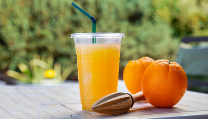 disposable plastic glass of cold freshly squeezed orange juice with wooden juicer on table outdoors. Fresh drink concept