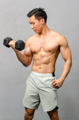 Asian strong handsome fit young male muscular shirtless fitness model athlete in sporty shorts standing posing lifting weight metal dumbbell showing biceps triceps muscle in gym on gray background