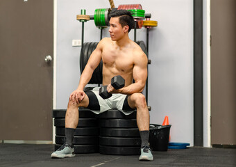 Millennial Asian fit strong handsome shirtless muscular male fitness model athlete in shorts sitting on barbell weights lifting dumbbell do biceps triceps working out training exercising alone in gym