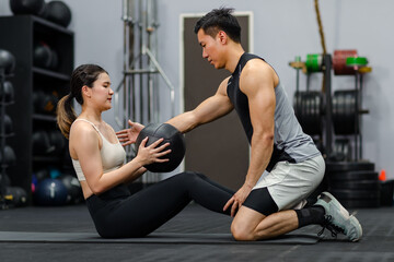 Fototapeta na wymiar Asian strong young male muscular fitness trainer helping female athlete model in sexy sport bra and legging lifting working out with heavy leather ball workout on floor exercise training in gym