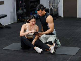 Obraz premium Asian strong young shirtless male muscular fitness model in sporty shorts sitting smiling holding showing tablet screen to female athlete in sexy sport bra while taking break after working out in gym