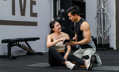 Fototapeta na wymiar Asian strong young shirtless male muscular fitness model in sporty shorts sitting smiling holding showing tablet screen to female athlete in sexy sport bra while taking break after working out in gym