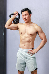 Fototapeta na wymiar Asian strong handsome young male muscular shirtless fitness model in sporty shorts standing smiling holding lifting metal kettlebell dumbbell showing biceps triceps muscle in gym on gray background