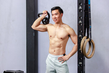 Asian strong handsome young male muscular shirtless fitness model in sporty shorts standing smiling holding lifting metal kettlebell dumbbell showing biceps triceps muscle in gym on gray background