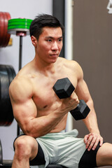 Naklejka na ściany i meble Millennial Asian fit strong handsome shirtless muscular male fitness model athlete in shorts sitting on barbell weights lifting dumbbell do biceps triceps working out training exercising alone in gym