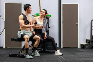 Fototapeta na wymiar Asian strong young male muscular shirtless fitness model in sporty shorts and female athlete in sport bra sitting smiling taking break holding water bottle talking together in gym
