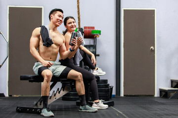 Fototapeta na wymiar Asian strong young male muscular shirtless fitness model in sporty shorts and female athlete in sport bra sitting smiling taking break holding water bottle talking together in gym