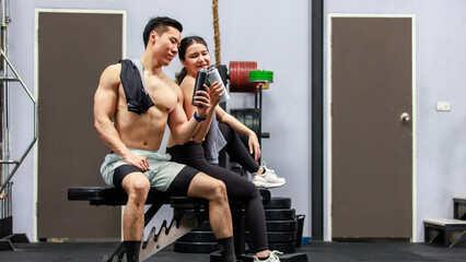 Obraz premium Asian strong young male muscular shirtless fitness model in sporty shorts and female athlete in sport bra sitting smiling taking break holding water bottle talking together in gym