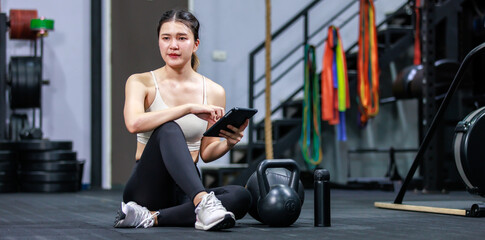 Fototapeta na wymiar Millennial Asian strong young fit female muscular fitness athlete model in sportswear sport bra and legging sitting on floor taking break from working out checking message on touchscreen smartphone
