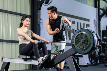 Asian strong young fit male trainer helping teaching female muscular fitness athlete model in sexy sport bra and legging sitting using cable pulling machine working out training exercising in gym