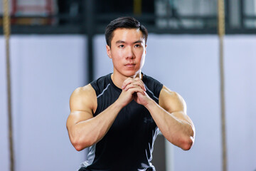 Millennial Asian strong young fit male muscular fitness athlete model in sleeveless shirt and sporty shorts training exercising squatting on wooden box alone in gym full of exercise equipment