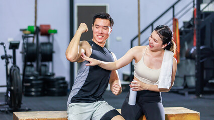 Asian strong young fit male muscular fitness athlete model showing off bicep muscle to female friend sitting take break drink water wipe sweat on wood box smiling hold thumb up complimenting in gym