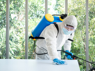 Asian worker man cleaning a white desk protection disease from Covid19 or bacteria while wearing...
