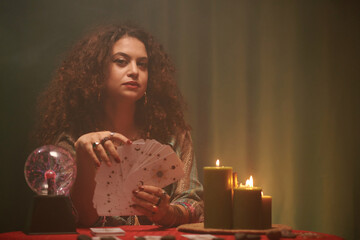 Portrait of fortune teller with tarot cards sitting at table with burning candles and crystal ball