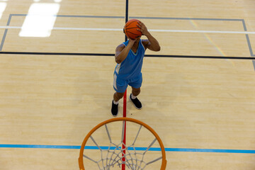 Biracial male basketball player wearing blue sports clothes and shooting basketball at gym