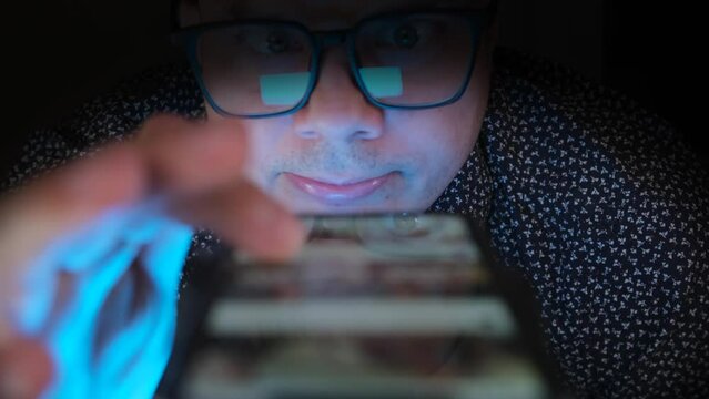 Funny inadequate unshaven man who has lost touch with reality from long stay in virtual space, both hands scrolls feed in social networks and online video platforms, close up.