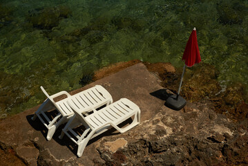Titolo: On the tip of a rocky cliff, two comfortable white deckchairs and a red umbrella, are we ready for a swim?
