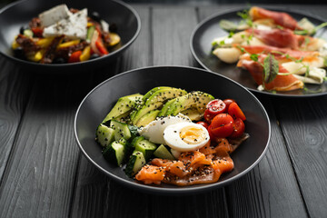 Salmon salad with tomato, cucumber, avocado, egg and sesame seeds on a plate on a black wooden table with other dishes for lunch