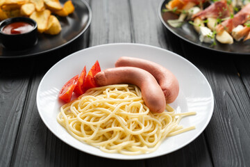 Spaghetti with boiled sausages and pieces of chopped tomato on a plate on a black wooden background