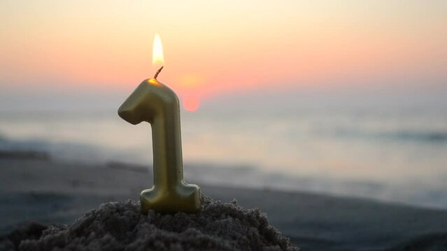 Numeral one from yellow gold color paraffin candle wax burns on sandy beach of sea shore at dawn sunset close-up. Paraffin candle burning near sea. Concept holiday Birthday New Year party celebrating