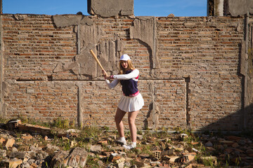 Young, beautiful, blonde, young woman playing baseball dressed in her baseball kit. Woman hits ball with baseball bat, background brick wall in a ruined building. Concept sports and olympic games.