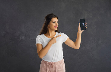 Smiling young Hispanic woman on black studio background show mockup screen on modern smartphone. Happy millennial Latin girl demonstrate copy space good deal sale on cellphone. Technology concept.