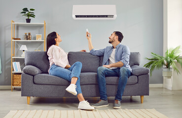 Young happy man and woman turning on air conditioner sitting on sofa at home. Smiling couple of homeowners enjoying cool conditioned air using remote resting on couch together in living room. - obrazy, fototapety, plakaty