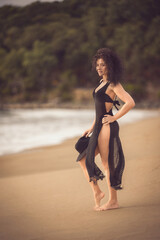 Fototapeta premium Captivating female model with curly hair and a slender sexual body poses elegantly in a chic black beach dress and hat, showcasing her attractive physique at the tropical beach.
