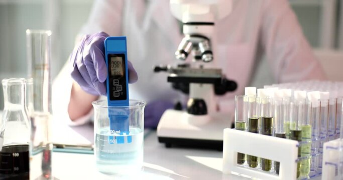Laboratory scientist in rubber gloves measures pH level of transparent chemical liquid. Sterileable with microscope and test tubes. PH meter