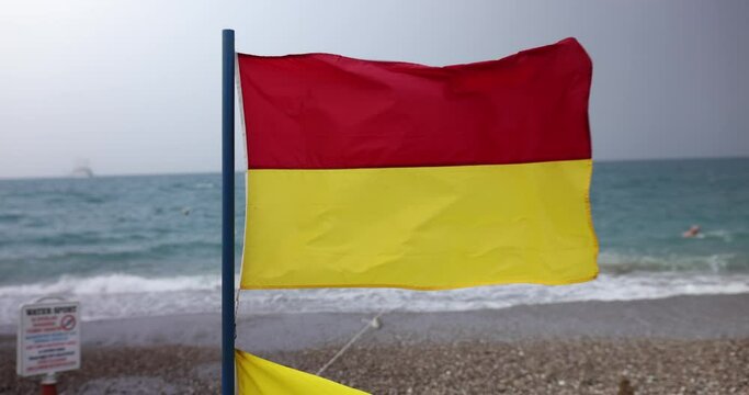 Red and yellow rescue flag flutters in strong sea breeze on pebble beach. Flag signaling of stormy weather and warning of swimming in sea