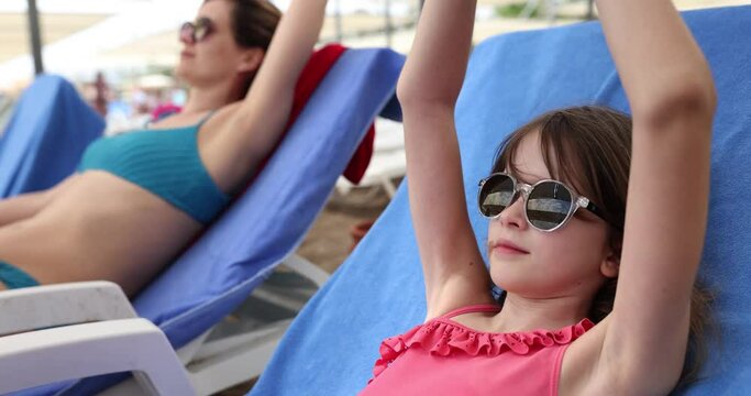 Junior schoolgirl in sunglasses lies on beach sun lounger near relaxing mother. Teen girl throws hands behind head sunbathing. Holidays in exotic resort