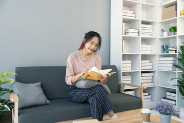 Fototapeta na wymiar Young Asian woman reading a book at her home sitting on the sofa In the living room