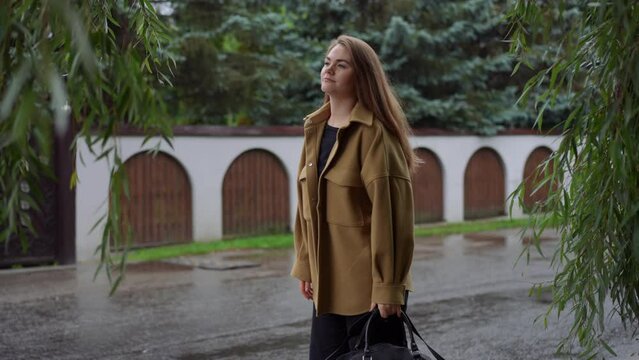 Side view portrait of young Caucasian brunette woman with bag hitchhiking standing on overcast street. Medium shot of charming beautiful lady with luggage catching transport on rainy day outdoors