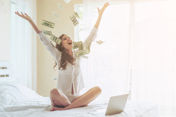 Beautiful woman sitting on the bed in front of her laptop , sprinkle money on the bed with a happy smile  Because she profits from investing in online stocks - start up online business concept