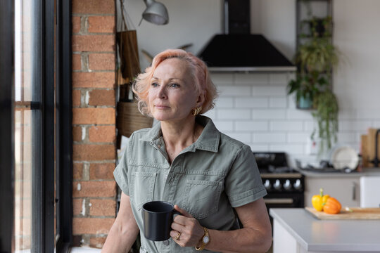 Senior Woman At Home Alone Looking Out Of Window Thinking And Having A Hot Drink